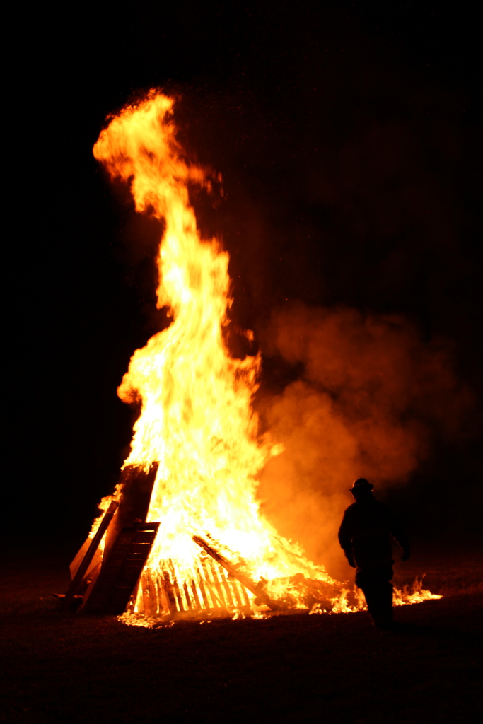 Series of photos from the West Carroll Homecoming Bonfire held at night on school grounds. Students and staff gather with friends around a large bonfire, pose for group photos, and enjoy the Homecoming celebration. West Carroll cheerleaders perform near the fire and serve refreshments, while additional photos show the bonfire being prepared and lit with safety personnel nearby.