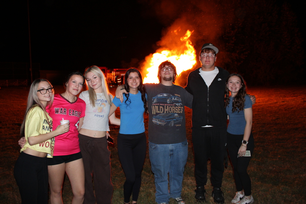 Series of photos from the West Carroll Homecoming Bonfire held at night on school grounds. Students and staff gather with friends around a large bonfire, pose for group photos, and enjoy the Homecoming celebration. West Carroll cheerleaders perform near the fire and serve refreshments, while additional photos show the bonfire being prepared and lit with safety personnel nearby.