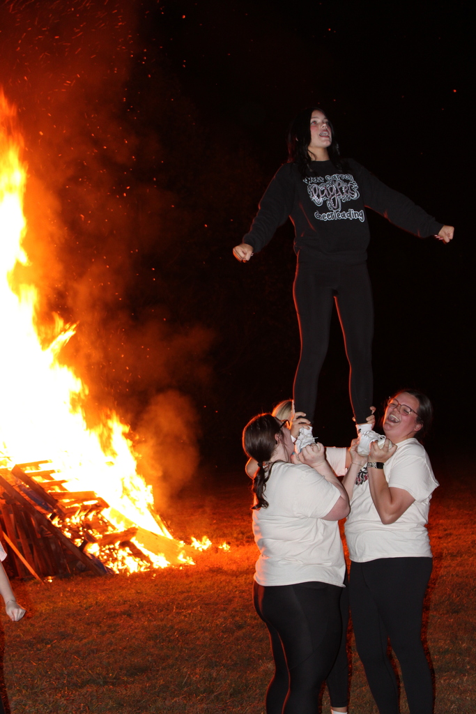 Series of photos from the West Carroll Homecoming Bonfire held at night on school grounds. Students and staff gather with friends around a large bonfire, pose for group photos, and enjoy the Homecoming celebration. West Carroll cheerleaders perform near the fire and serve refreshments, while additional photos show the bonfire being prepared and lit with safety personnel nearby.