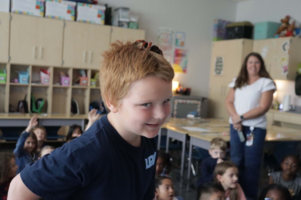Boy with red hair smiling with a tarantula spider on his head. Children, a teacher, and cabinets are visible behind him