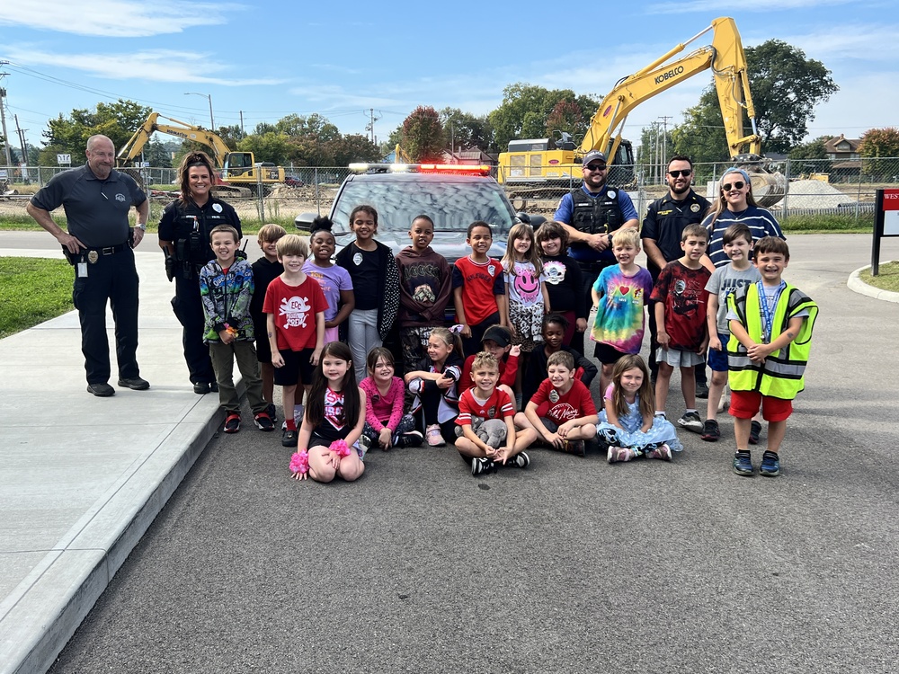Group of children, some sitting and some standing, in front of a police cruiser. Four police officers and one teacher can be seen in the back. A construction vehicle and blue sky is visible in the background.
