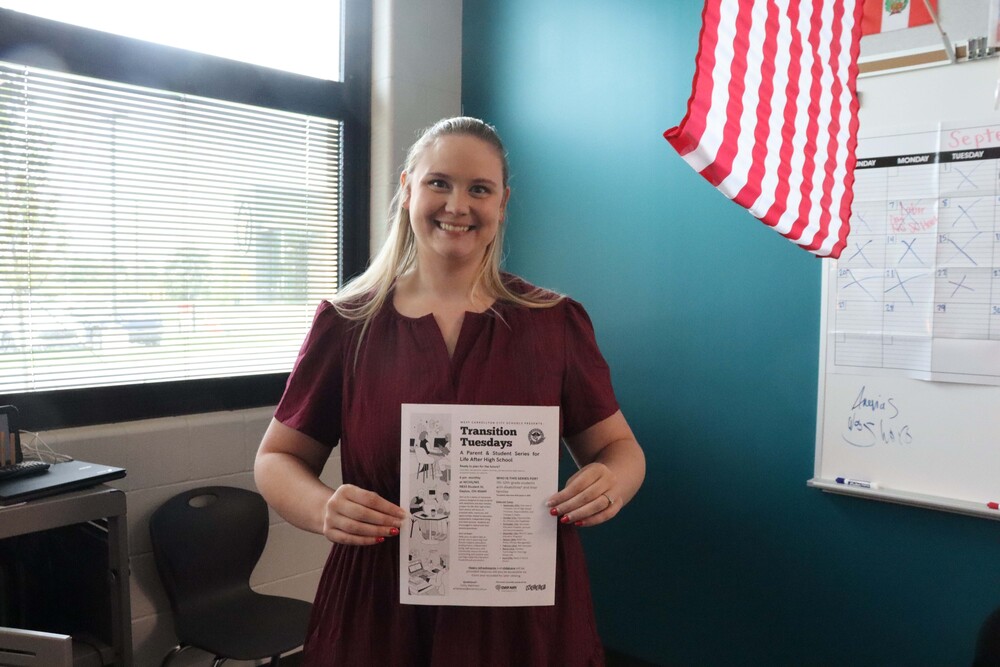 A teacher smiling and holding a flier with the test "transition tuesdays". A flag, calendar, window and green wall are present in the background.