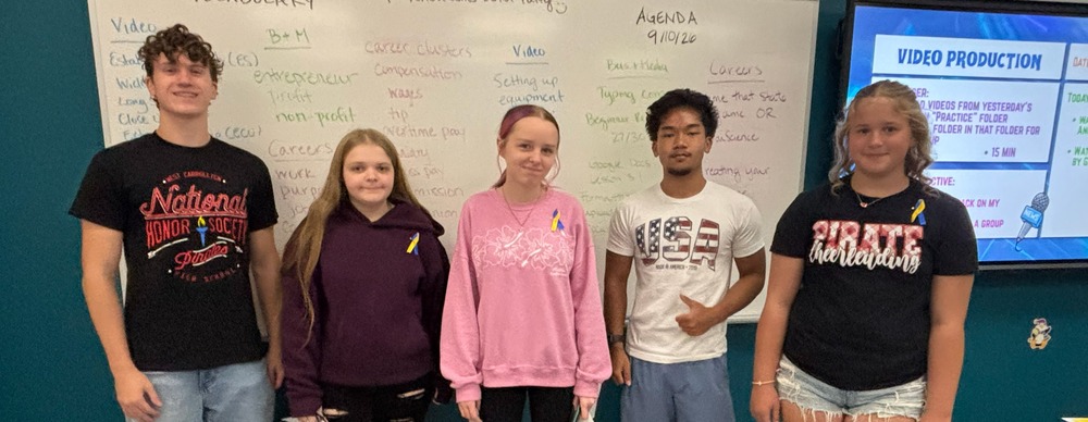 Five young adults stand in a classroom with a whiteboard in the background