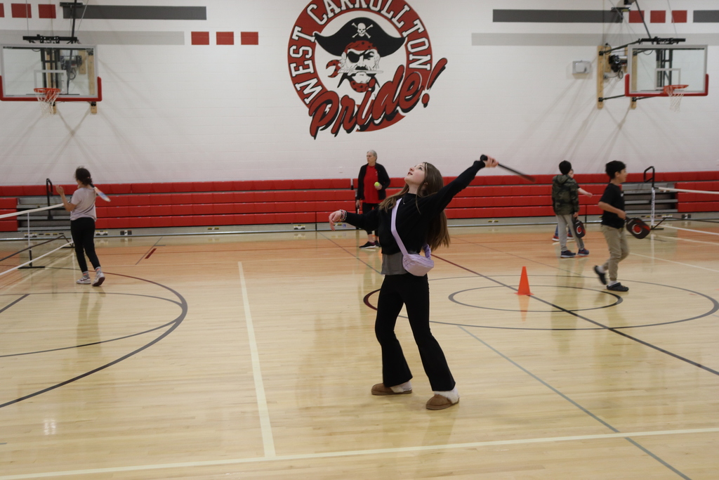 A group of individuals are playing pickleball indoors. One person swings a racket while others stand around.