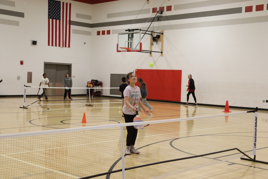 People playing pickleball in a gym with a red wall, American flag, and orange cones on the floor.