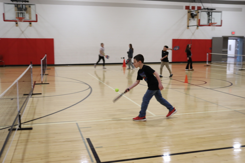 Multiple people playpickleball in a gym with orange cones. A boy in black and blue swings a racket.