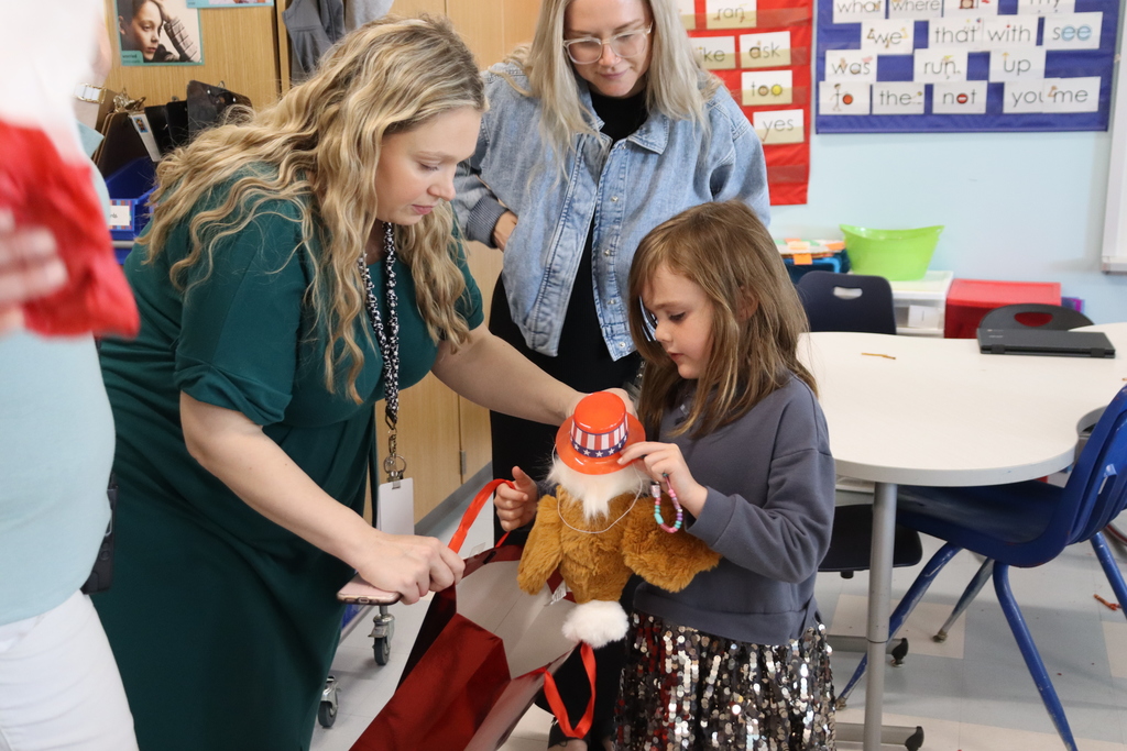 A woman assists a young girl holding a stuffed toy while standing in a classroom with tables and chairs.