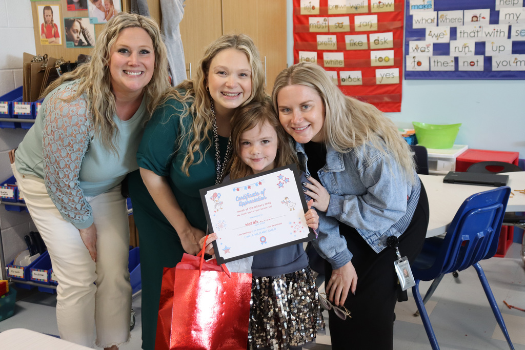 Three women and a young girl stand in a classroom. The girl holds a certificate.