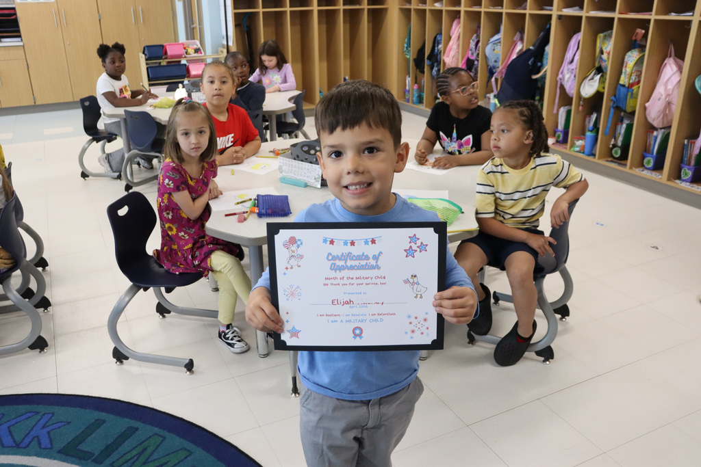 A child holds a certificate in a classroom. Other children sit at tables behind him.