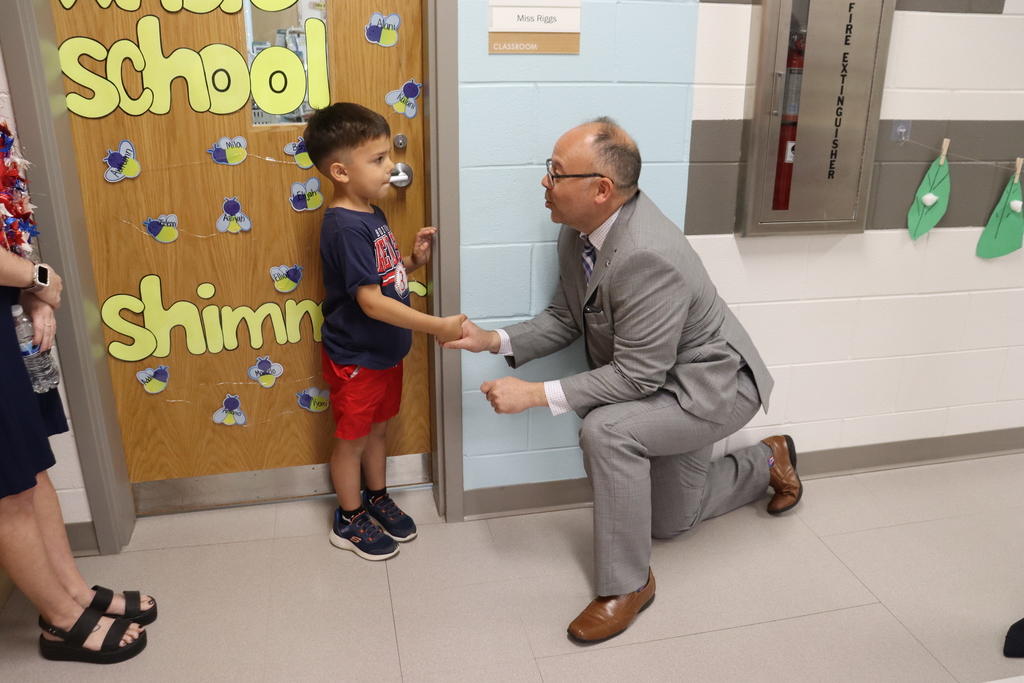 A man in a suit kneels to greet a young boy at a classroom door. The door is decorated with colorful drawings.