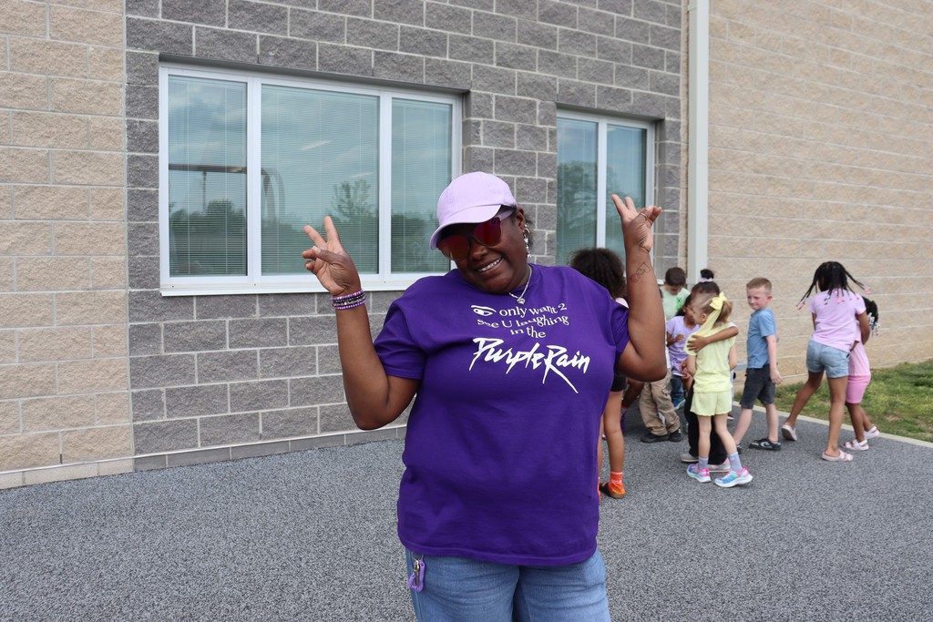 A person in a purple shirt and hat makes a peace sign in front of a brick building.