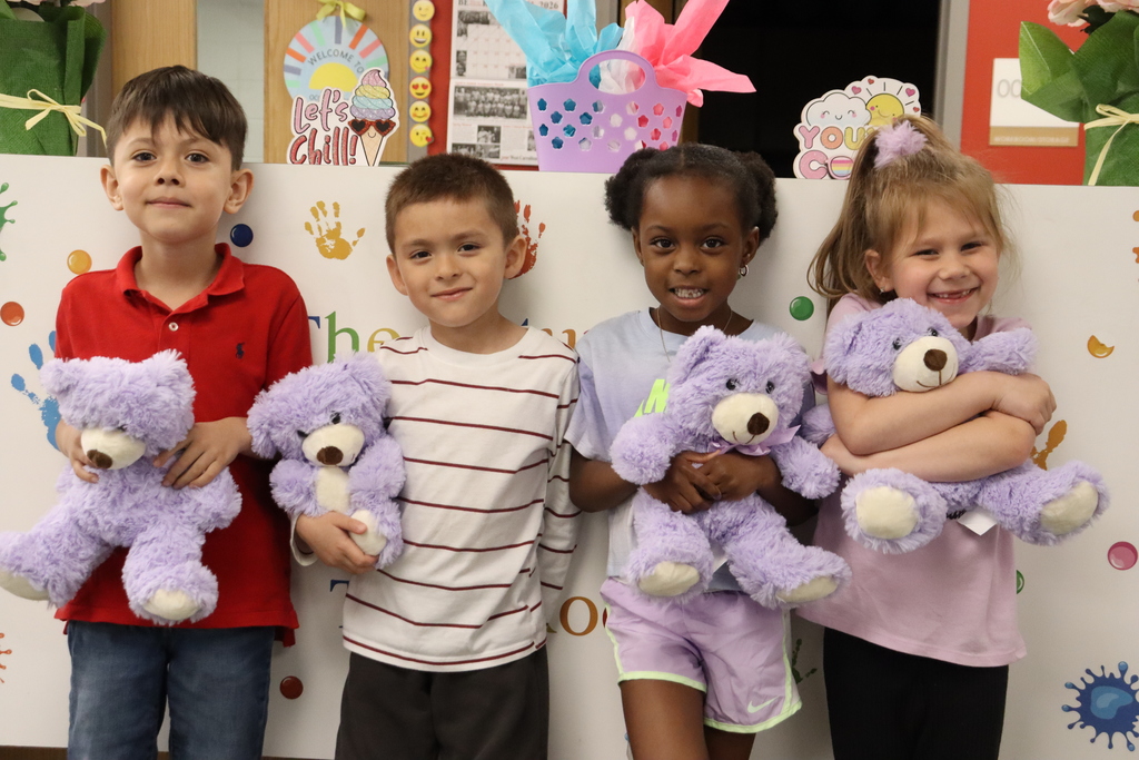 Four children in a classroom hold purple stuffed animals. 