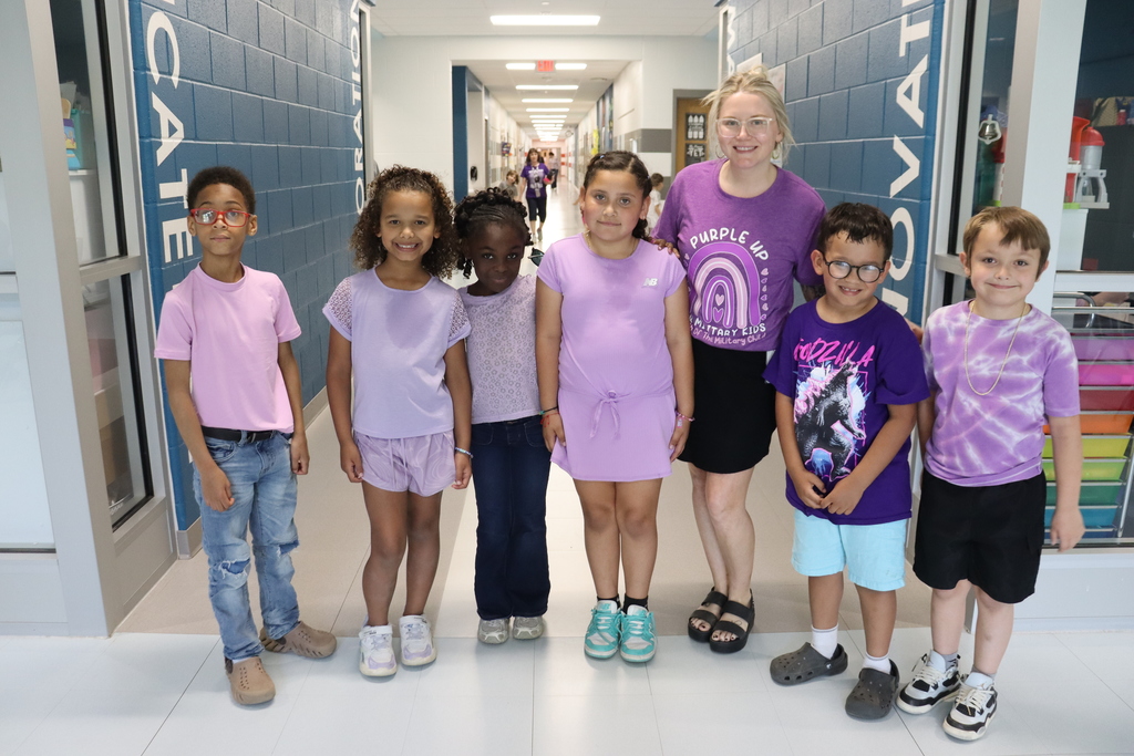 A group of six children and a woman stand in a hallway. They wear matching purple T-shirts.