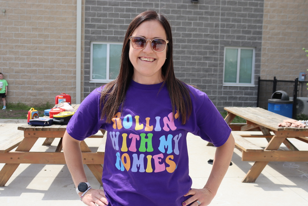 A woman stands with hands on hips, smiling. Behind her, a brick building, picnic tables, and a trash bin.