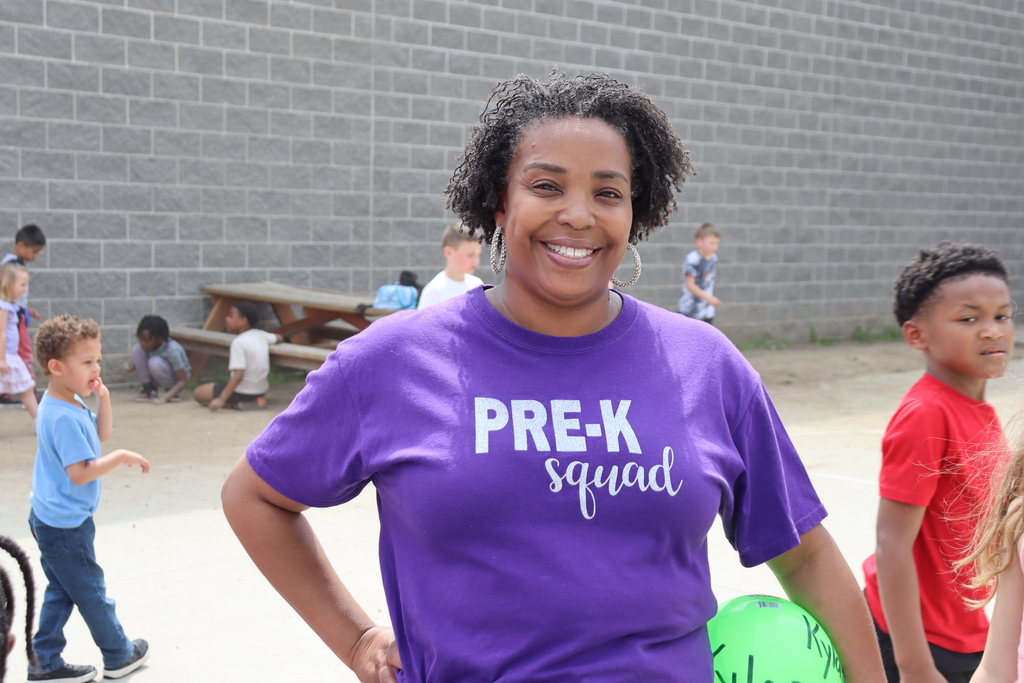 A woman with curly hair wearing a purple shirt with "PRE-K squad" smiles. Behind her, children play, one holding a ball.