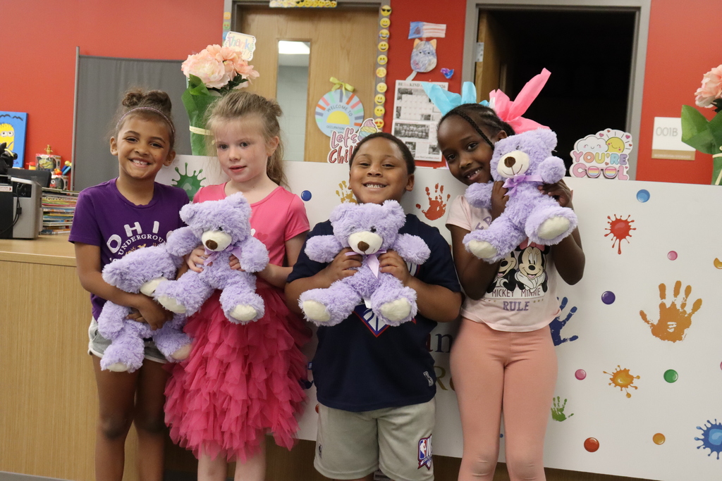 Four young children stand together, each holding a purple teddy bear. They pose against a colorful wall with handprints.