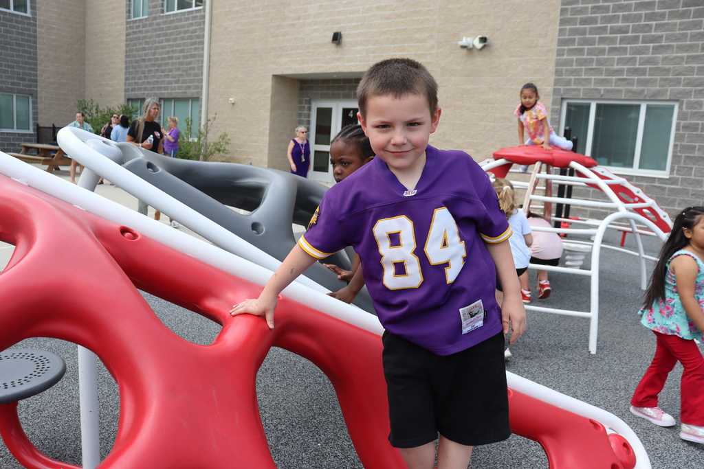 A boy in a purple shirt with number 84 stands near a red slide at a playground.