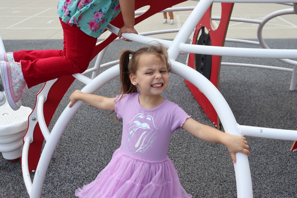 A young girl in a purple dress smiles at a playground with another child behind her.