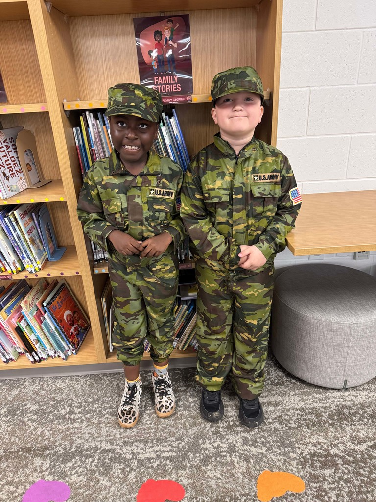 Two children in camouflage uniforms standing in a library with bookshelves in the background.