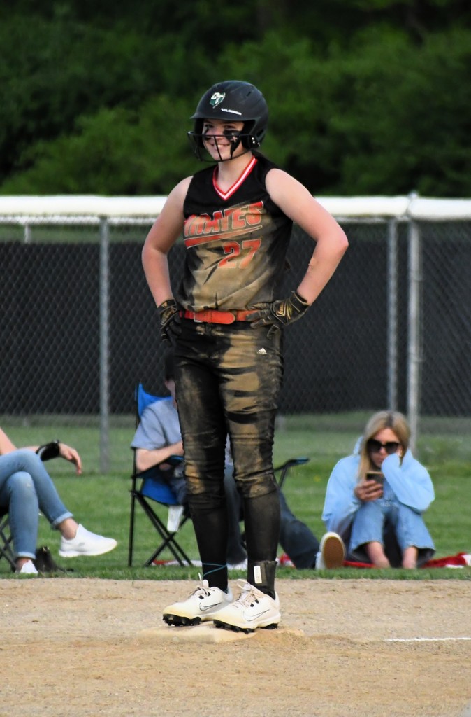 Player in black and red uniform stands on the field, looking away. Behind, a fence, trees, and spectators.
