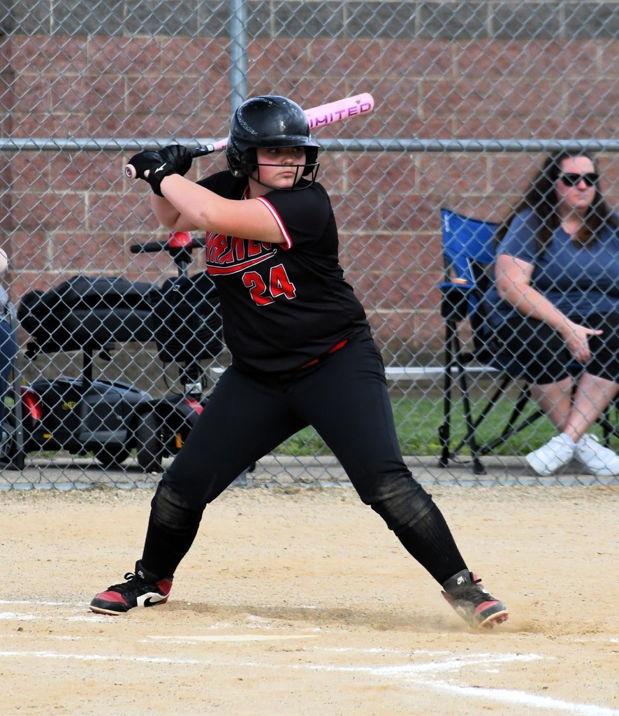 A softball player in a black uniform, ready to hit, stands at home plate, with a fence and spectators in the background.