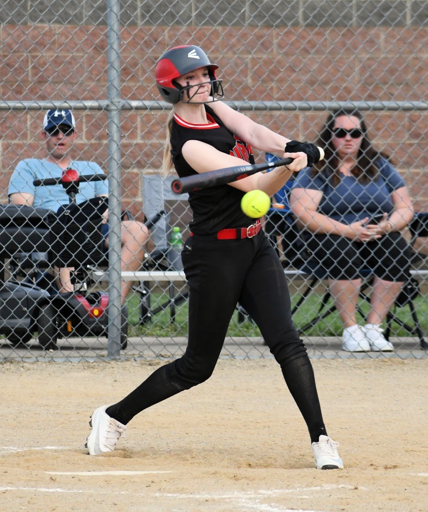 A softball player swings her bat at a pitch. Behind her, two spectators watch. In the background, a metal fence.
