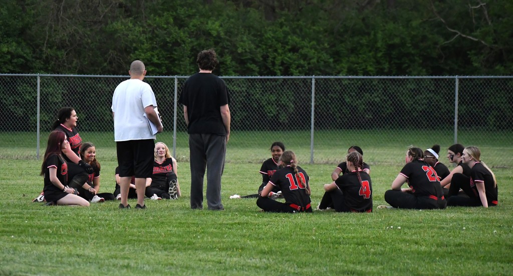 A group of athletes wearing black uniforms sit on grass. A coach stands nearby, holding a clipboard.