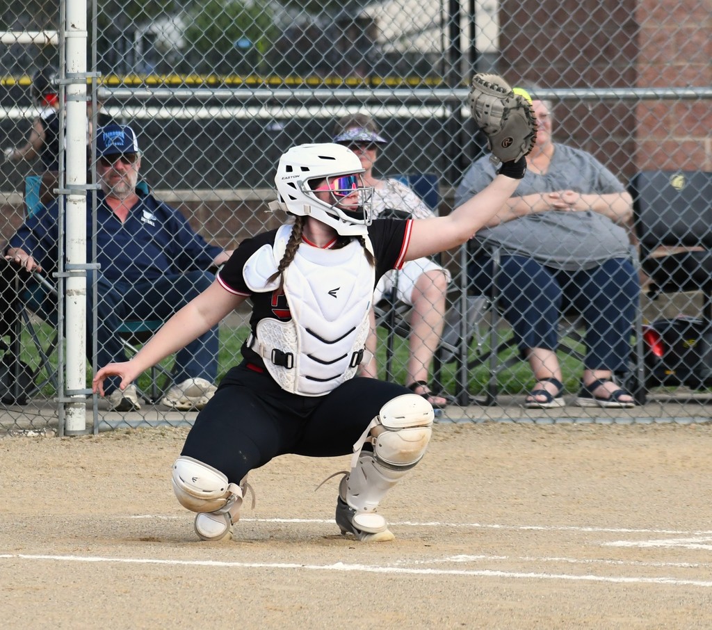 Woman in a white helmet crouches with knees bent, arm extended at a softball game. Behind her, spectators sit behind a fence.