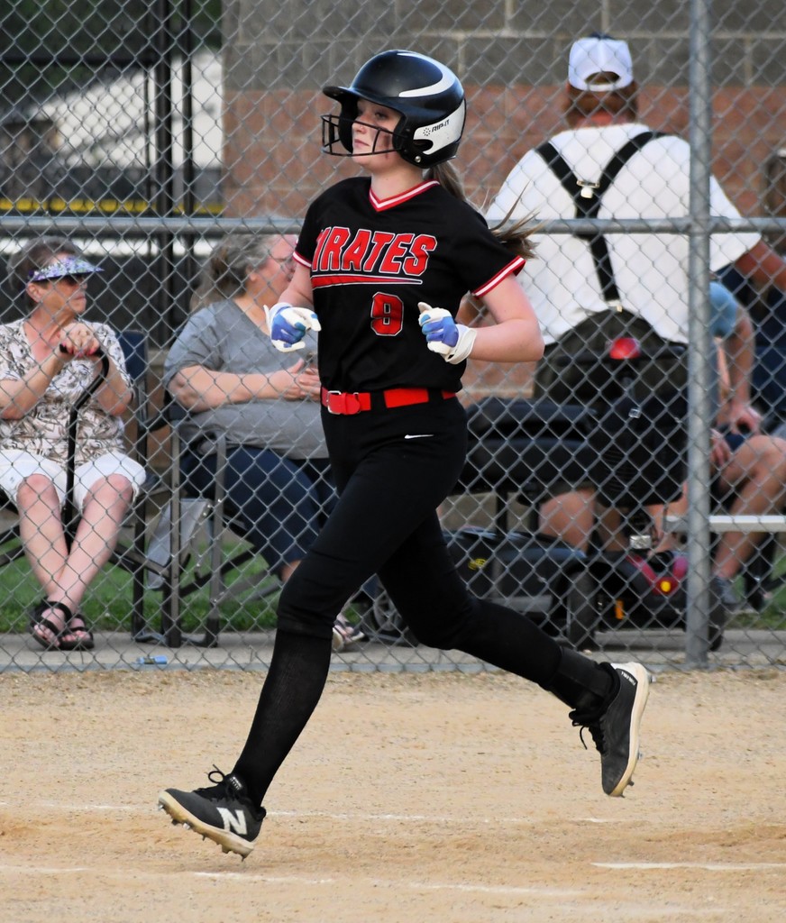 Player in black jersey and helmet running toward base, spectators behind chain-link fence.