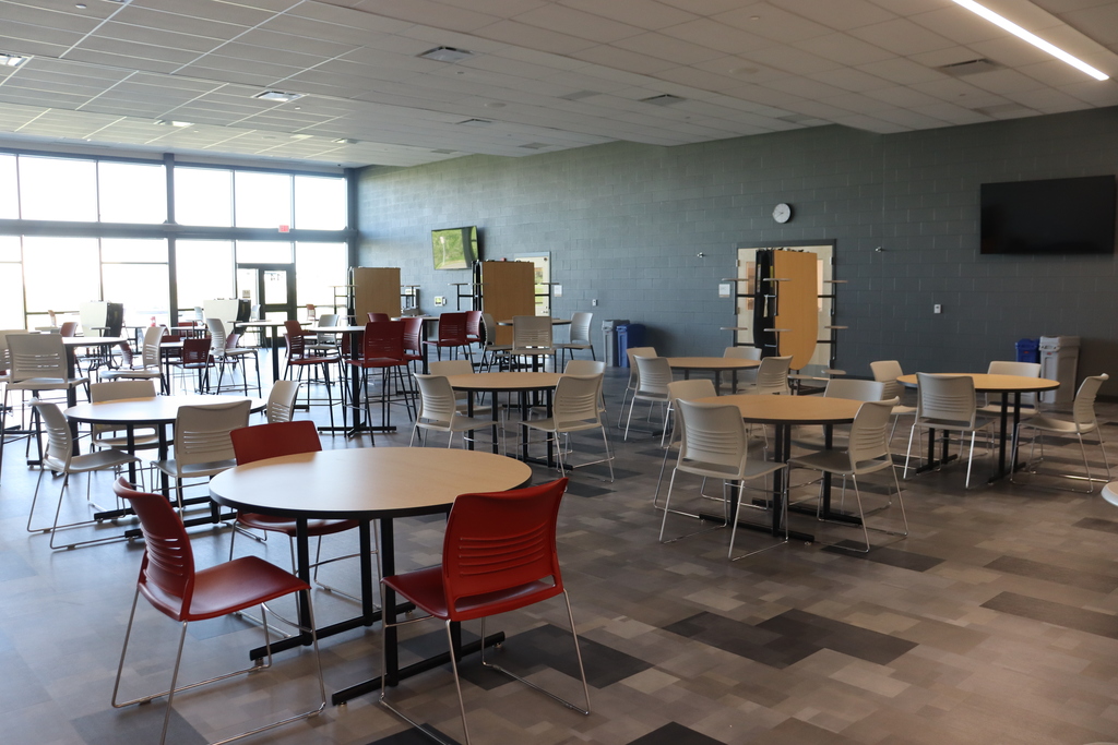 Empty dining hall with round tables and chairs. Light gray walls, windows, and a television. Floor has a geometric pattern.