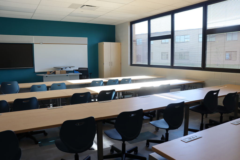 An empty classroom with rows of desks and chairs. A whiteboard is mounted on the wall, and large windows are on the right side.