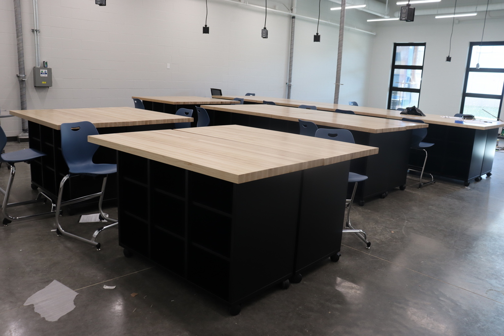 An empty room with gray floors and white walls. Several wooden desks with blue chairs are in rows.