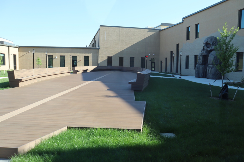 A courtyard with benches on a wooden deck. A grassy area, a large sculpture, and a building.