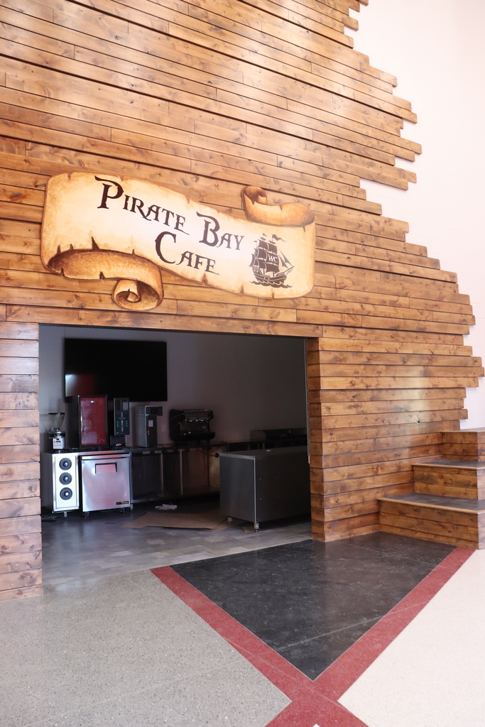 Wooden wall with a pirate-themed sign reading "Pirate Bay Cafe". Entrance leads to a kitchen area with appliances.
