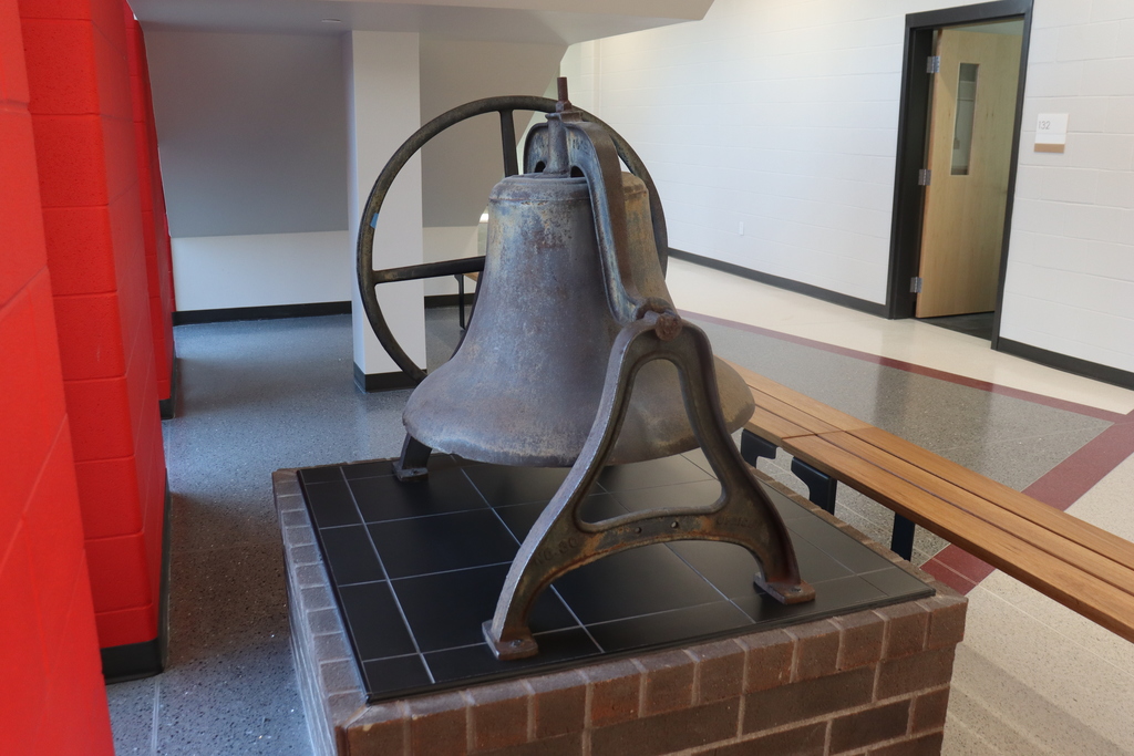A large bell on a brick base with a metal frame is displayed in a hallway. Behind it, there are red columns and a door.