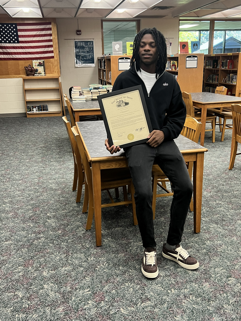 A young person in a library, holding a certificate, wearing a jacket and sneakers, sitting at a table.