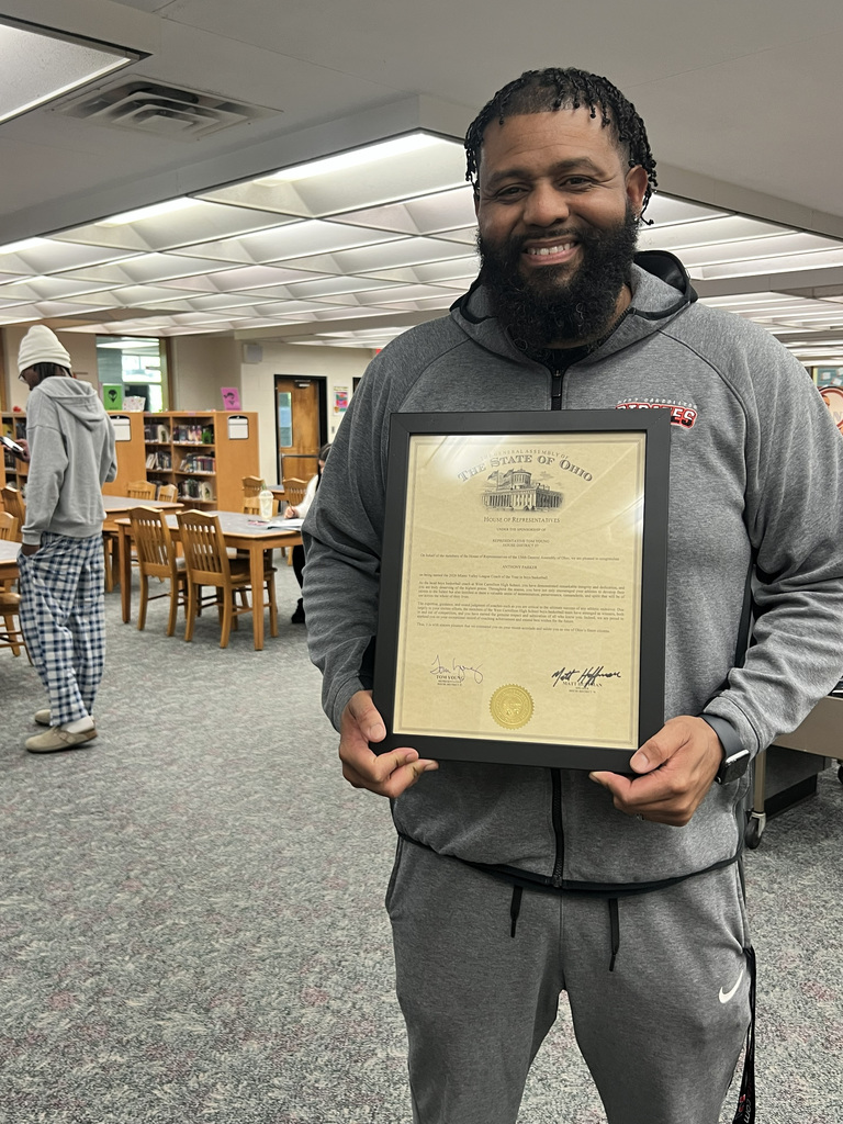 Man in gray sweatshirt holding a framed certificate in a library with chairs and tables in the background.