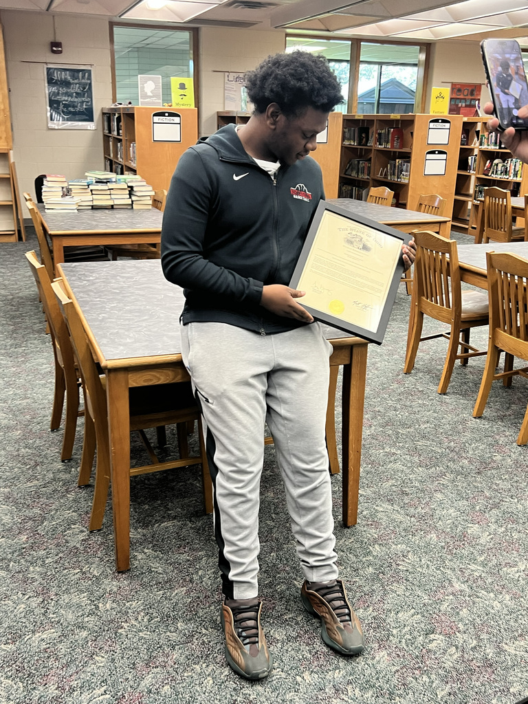 Man standing in a library, holding a framed certificate, with wooden tables and chairs in the background.