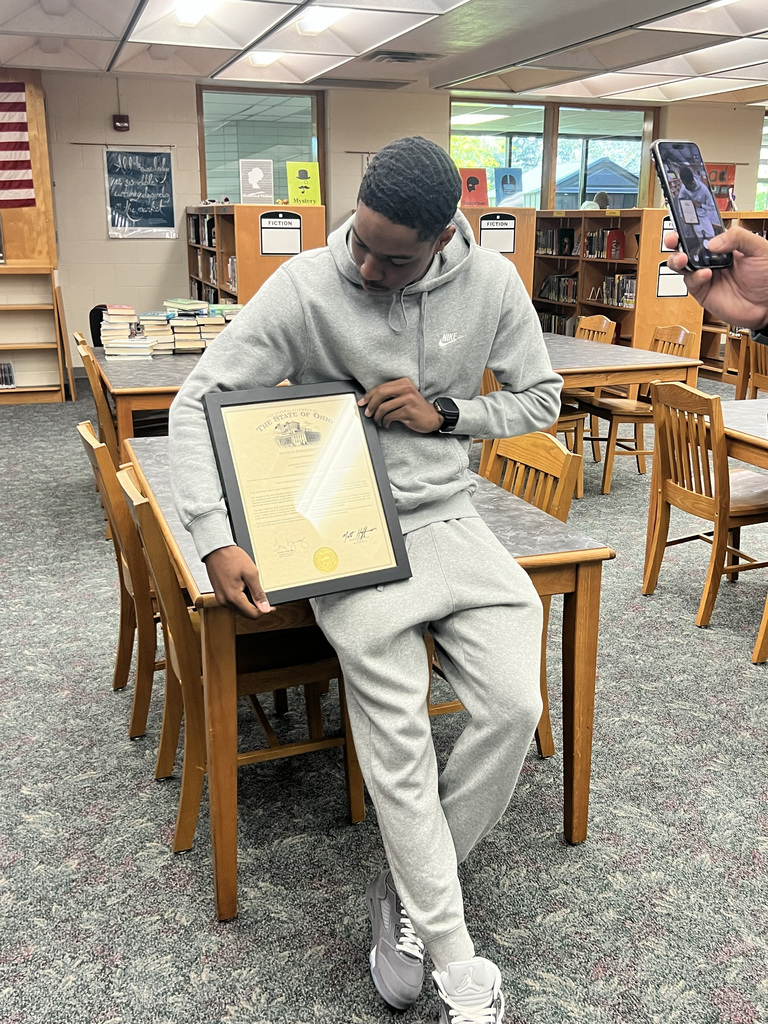 A person holds a framed certificate while sitting at a table in a library. Behind them, chairs and tables are visible.