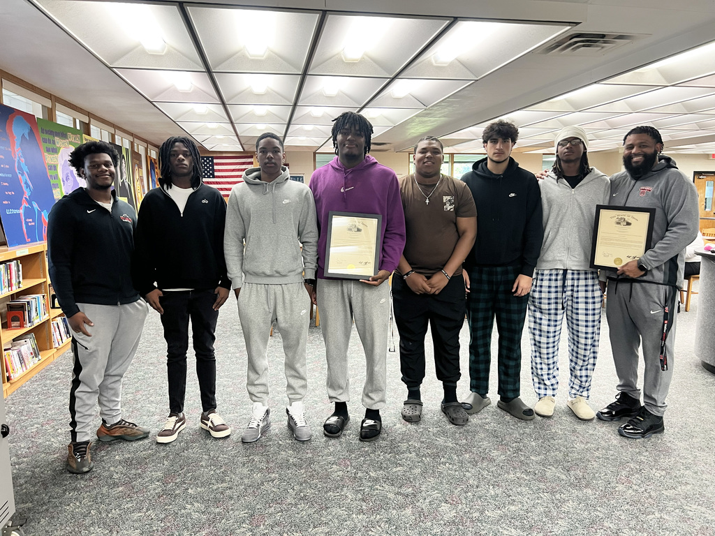 Eight men stand in a row in a library, holding certificates. One wears a purple shirt, and others wear black, white, and gray clothes. Behind them, shelves with books are visible.