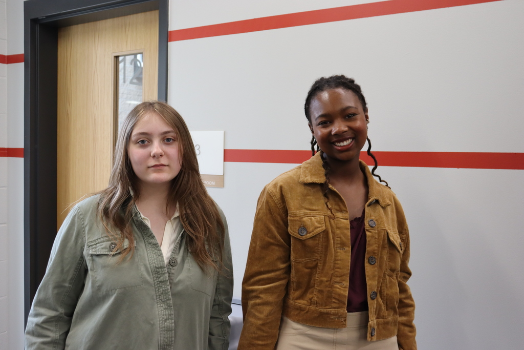 Two women stand next to each other in a room with white and red-striped walls.
