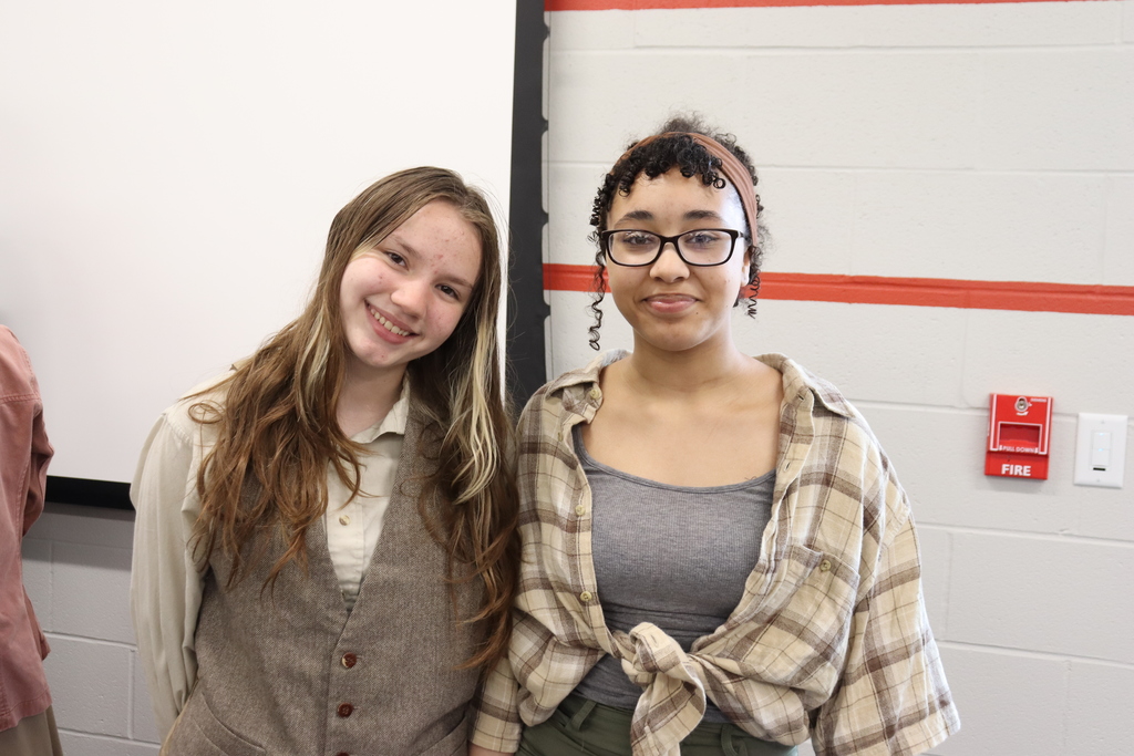 Two women stand in front of a white wall with a red stripe, both smiling. One wears a gray top and plaid jacket, the other a gray vest over a white shirt.