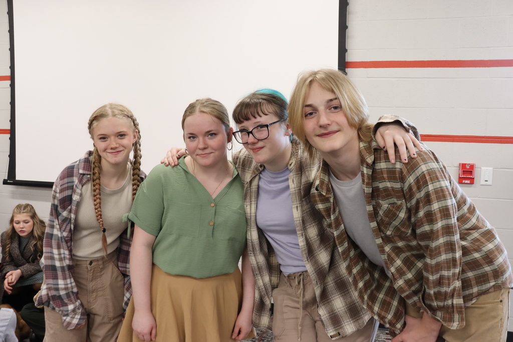 Four individuals in a classroom, smiling for a photo. One wears glasses, another has braided hair. Background shows a whiteboard and people seated.