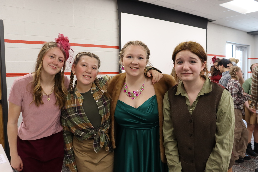 Four young women stand together, smiling, with two wearing vintage-style dresses. A red line marks the wall behind them.