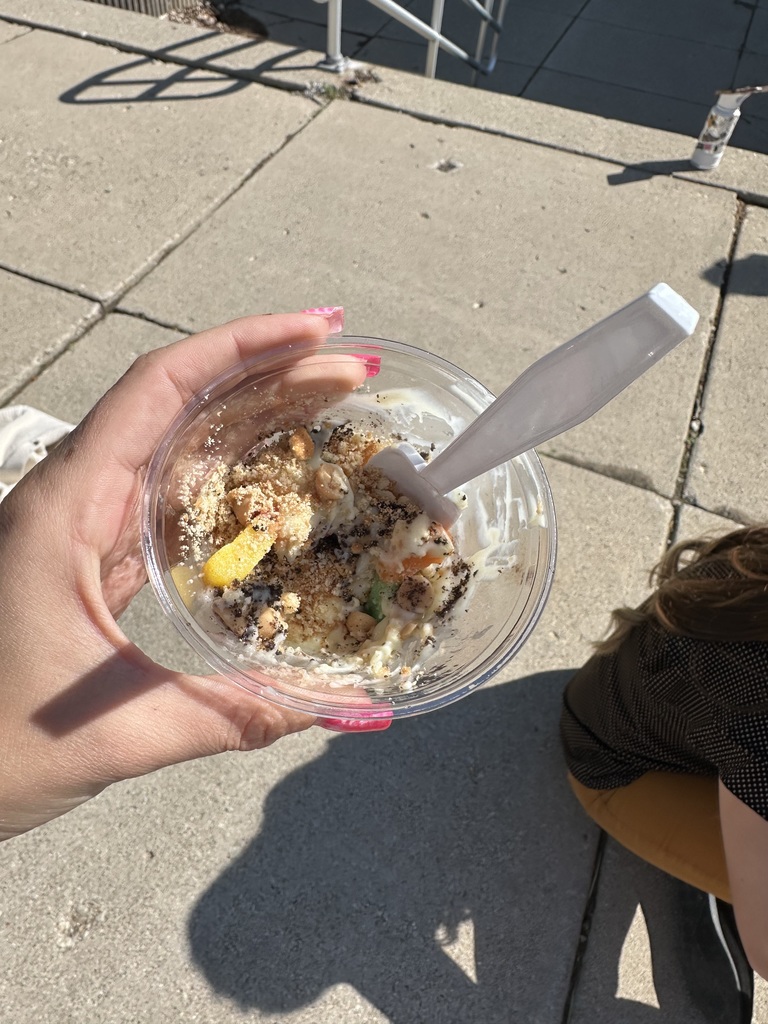 Person holds a bowl of food with a spoon on a sidewalk with a trash can nearby.