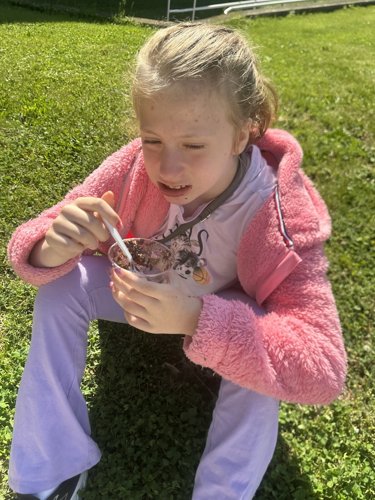 A child wearing a pink jacket and purple pants sits on grass, eating from a bowl.
