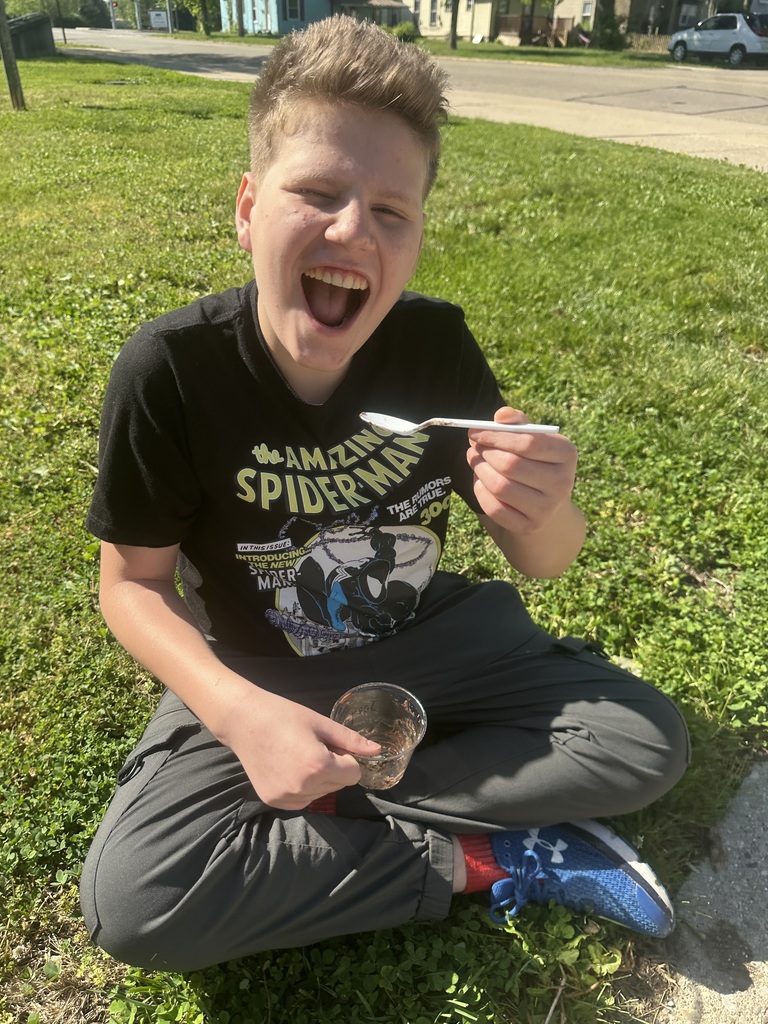 A boy wearing a black t-shirt, gray pants, and sneakers is sitting on grass, holding a spoon with food and laughing.