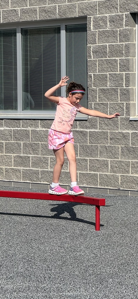 A girl balances on a red beam outside a building with a brick wall and windows.