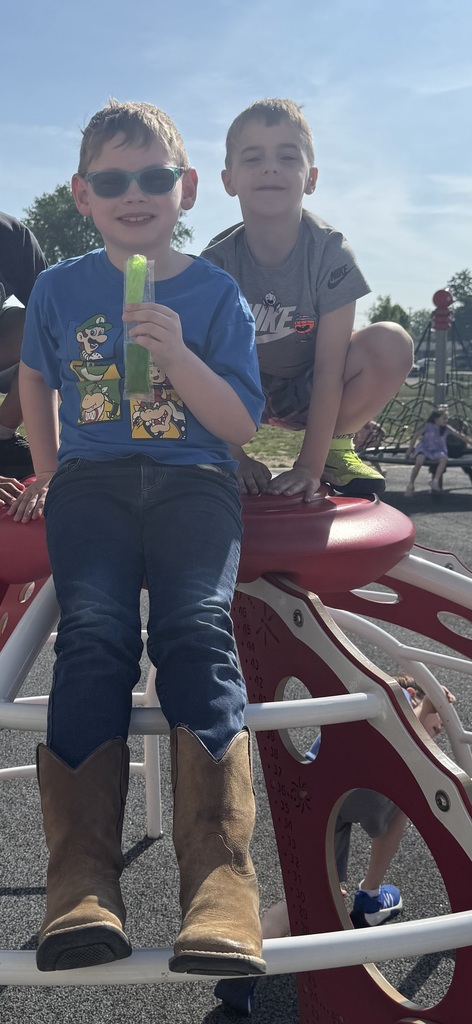 Two boys on the playground. One holds a green object and sits on a curved red structure. Another boy stands behind him on a smaller red structure.