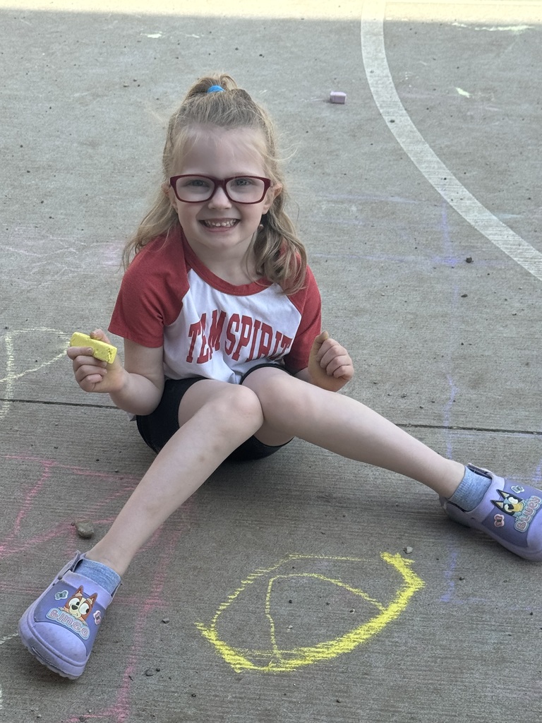 A young girl wearing glasses  is sitting on the ground with chalk.